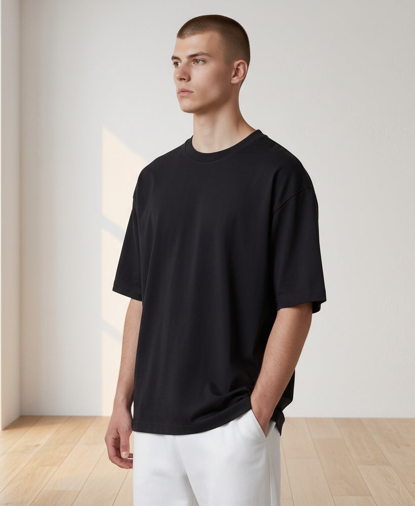A man wearing an oversized black short-sleeve crewneck t-shirt and white pants, standing in a minimalist room with light wood floors and off-white walls.